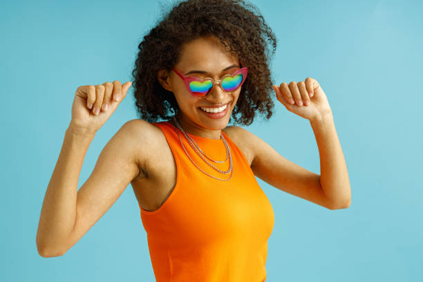 istock Happy woman in orange dress dancing with raised hands while standing over blue studio background 1654486402