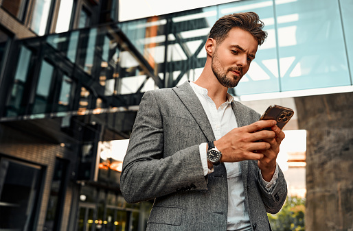 Business concept. Handsome attractive confident successful stylish serious bearded business man is holding a phone and texting while standing outside on the background of a business building.