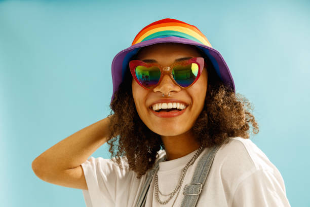 istock Smiling woman in rainbow cap and heart-shaped glasses looks camera over blue studio background 1654493607
