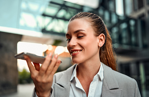 Portrait of a beautiful confident smiling adult professional woman standing against the background of a business center holding a phone and dictating a plan, reminders, questions in the application.