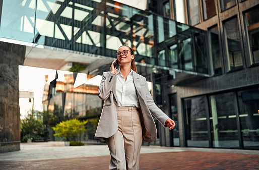 Attractive sincere happy confident adult professional woman in glasses and suit walking on the street near the business center of buildings and talking on the phone. Business lifestyle concept.
