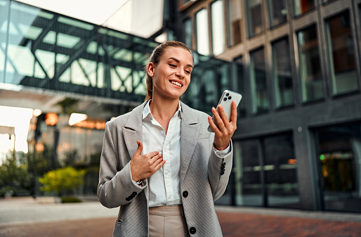 Portrait of an adult stylish elegant happy beautiful business woman in formal wear holding a phone and talking on video call on the street among business buildings. Corporate business call.