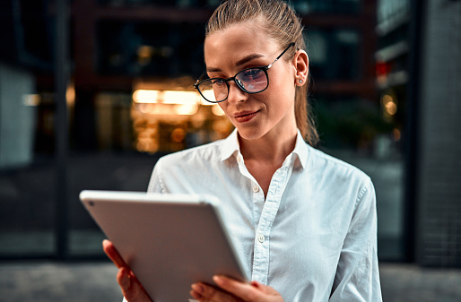 Beautiful adult modern smiling business woman in glasses holding a tablet and working on it. A professional office manager is standing on the street of a big city studying business finance analytics.