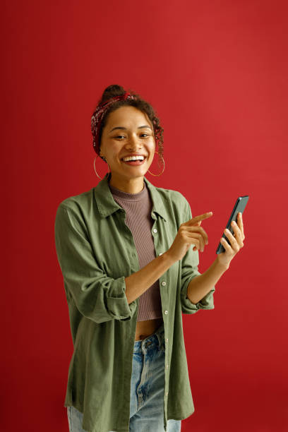 istock Smiling woman is pointing on mobile phone while standing on red studio background 1654523119