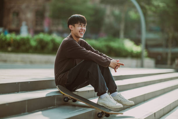 Full length portrait of a happy handsome young Asian man sitting near his skateboard sitting on the sidewalk during the day.