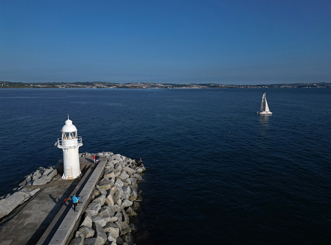 The drone camera gives a high angle view of Brixham Harbour Lighthouse and the Outer Harbour wall as a lone yacht heads out to sea early on a late, summer morning; in the background is the Torbay coastline. Brixham is both a major fishing port and a popular UK holiday and tourist destination.