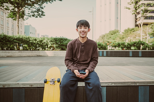 A young, happy Asian male traveler is enjoying his vacation, exploring the city with his skateboard, and using a mobile phone. Looking at camera
