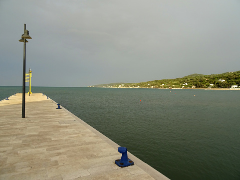 Rodi Garganico, Province of Foggia, Italy - 28 august 2023: one of the entrance piers to the tourist port and the eastern beach with the profile of the Gargano promontory up to the seaside village of San Menaio