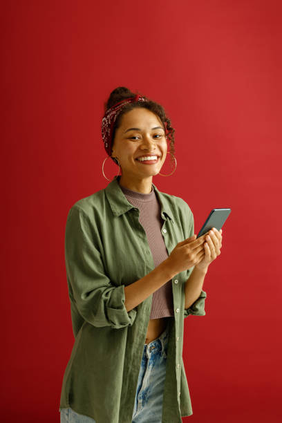 istock Pretty woman is using mobile phone while looks camera with smile on red studio background 1654545649
