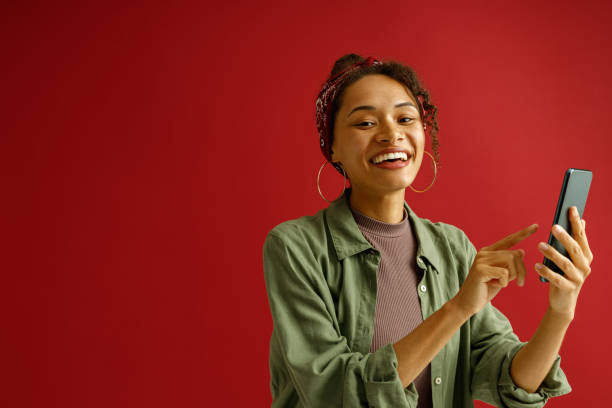 istock Smiling stylish woman is using mobile phone while standing on red studio background 1654546362
