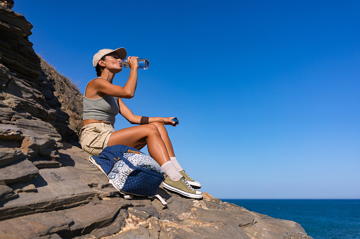 tourist girl with a backpack in the summer rests and drinks water sitting on top of a mountain