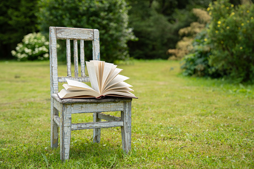 Open book on an old small childish chair with cracked paint on a green meadow in summer garden outdoors