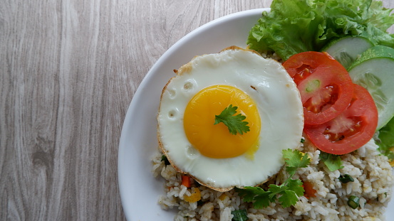 flat lay or top view of fried rice with egg and garnish with tomato, lettuce, cucumber and coriander leaves on a white plate with wood background.  Indonesian food.