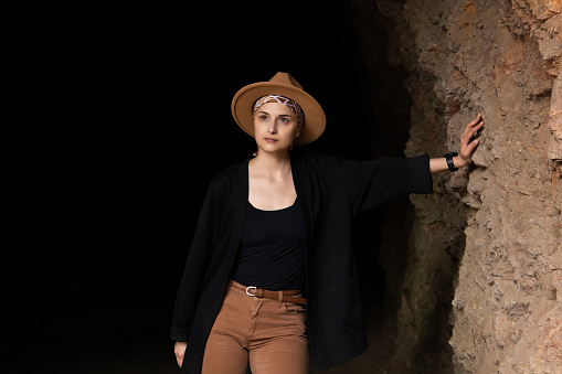 Woman archaeologist in a beige hat looks to the side, leans her hand against the wall of a dark cave.
