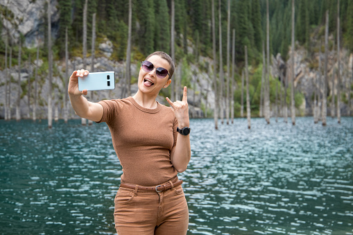 A young woman travels through the Tien Shan mountains of Central Asia, makes a cheerful selfie against the backdrop of the picturesque mountain lake Kaindy in the mountains of Kazakhstan.