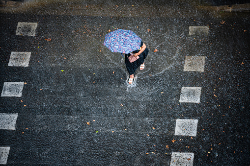 Aerial view of a woman with umbrella crossing street under heavy rain. High resolution 42Mp outdoors digital capture taken with SONY A7rII and Canon EF 70-200mm f/2.8L IS II USM Telephoto Zoom Lens