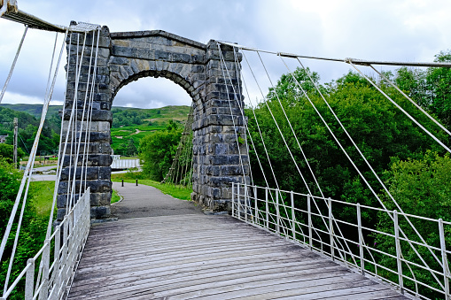 Fort Augustus, Scotland, July 6, 2023. Small suspension bridge over the River Caledonia (Called the Bridge of Oich) near Fort Augustus just off A82 in Scottish highlands. Set in wooded hillside valley. Summer day outdoors