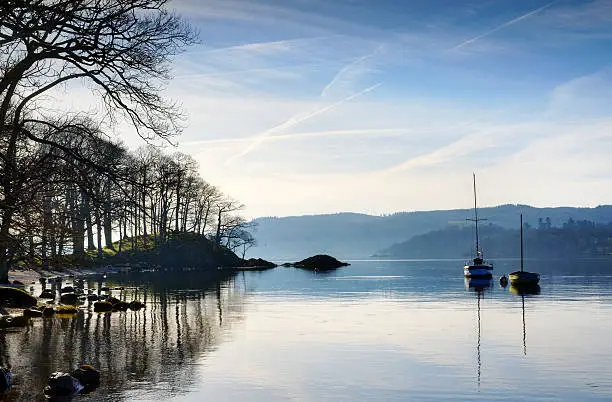 Beautiful view of a winter morning on Windermere in the English Lake District, with sunlight filtering through filigree branches and two boats calmly reflected in the water, set against a blue sky.