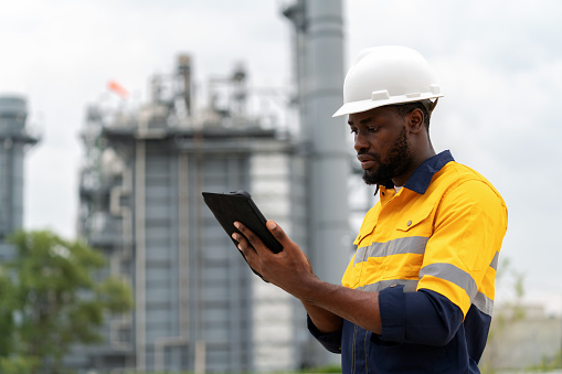 African America expert electrical engineer in safety suit and hardhat using a digital tablet to analyze plant data in the power plant factory. Ensuring the plant's efficiency and safety is paramount. Sustainable and social responsibility important to drive business.