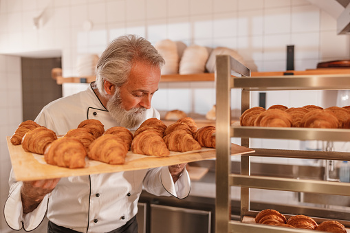 Three-quarter side shot with blurred background of an experienced mature Caucasian baker wearing a white cook uniform and a black apron while smelling and holding a tray full of freshly baked croissants and standing next to a baking tray trolley surrounded by all the bakery supplies.