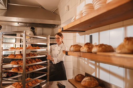 Three-quarter side shot with blurred right side of a young Caucasian female baker wearing a white chef's uniform and a black apron while pushing a baking tray trolley full of freshly baked croissants to to let them cool, surrounded by many varieties of breads.