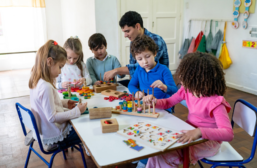 Diverse group of kids at school playing with wooden toys while male teacher supervises and interacts with them - Education concepts