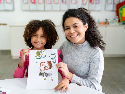 Latin American female teacher and her cute student holding a painting both looking very proud and happy at the camera - Education concepts