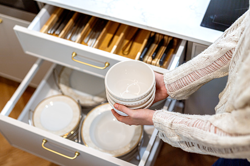 Woman organizing clean porcelain plates and bowls in kitchen drawer. Set of plates neatly organized. Tableware storage system in kitchen
