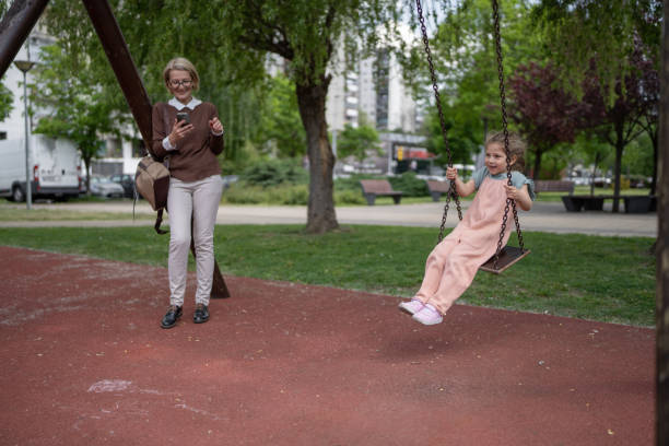 Young grandmother with her little granddaughter who is sitting on a swing in public park outdoors.