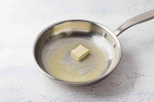 A piece of butter is melted in a frying pan on a light gray background. Bechamel sauce preparation step.