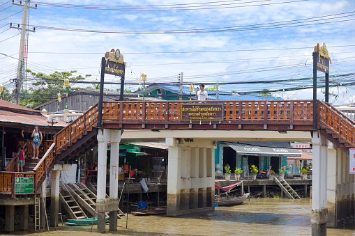 Bridge over river at Amphawa floating market area. Bridge has ornated railing and thai decor at both sides/ A few people are crossing the bridge. Some are on steps at left side. Behind bridge are old wooden houses with stores etc.