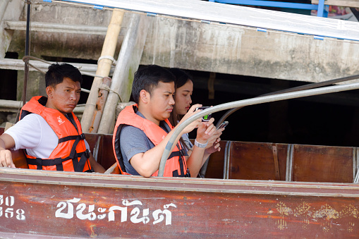 Capture of some thai people on  a tourboat in Amphawa. People are wearing life jackets for security. One man is using mobile phone. Capture from river and boat level. Scene is in area of old town and floating market below promenade