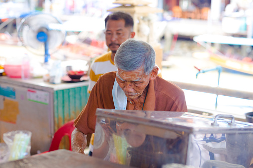 Portrait of thai senior market vendor with gray hair captured on promenade of floating market area in Amphawa. Man is is working concentrated and has a towel over shoulder. In background a  man is standing