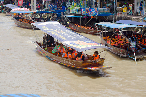 Tourboats on river in Amphawa between buildings at floating market area of old town. People in driving tourboat are wearing life jackets. Several tourboats are anchored along promenade