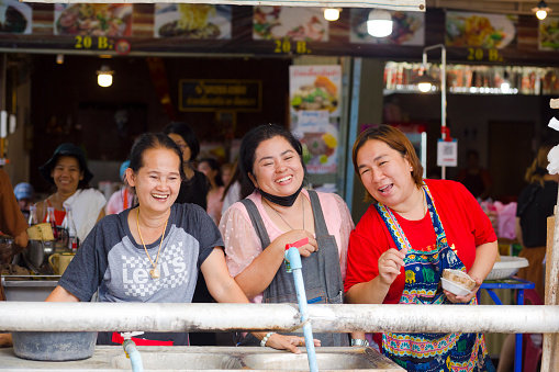 Capture of three happy laughing thai vendor women in Amphawa in area of old town and floating market. Women are standing at back of their market stall and are looking and communicating with people down in river. In background people and more market stalls are on prpomenade