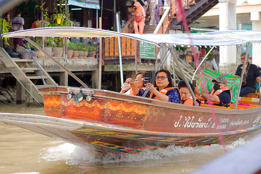 Passing by tourboat with tourists in Amphawa captured from boat on river in area of old town and floating market. In front of boat women are sitting. One is using mobile phone for video or photo. Behind  a man is sitting and wearing a protective face mask