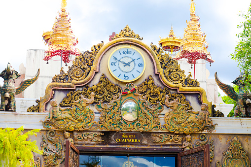 Detail of gate to เทวสถาน ชัยนครา Temple of Chamana in Amphawa with ornated golden clock. Around and below clock is artful relief. On wall are winged mystic animals. In background are golden stupas.