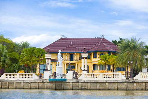 Old styled thai mansion and villa at river Mae Khlong in Amphawa seen from river. At waterfront are classical roman greek styled sculptures. In garden are palm trees