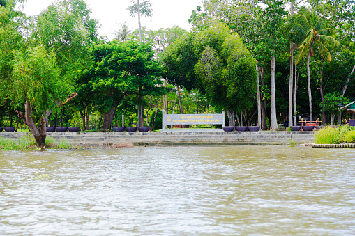 Waterfront of King Rama II Memorial Park in Amphawa with informational sign at river between trees. On ground are historical wooden thai buildings between trees