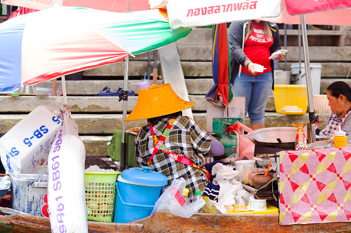 Rear view of thai senior woman cooking on a boat at floating market in Amphawa. Woman is wearing a straw hat. Another woman is at right side. Captured and seen from river and boat level. Behind woman and boat are steps to promenade. Scene is in old town and floating market area