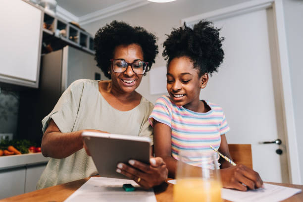 Mother using digital tablet helping her daughter with homework in the kitchen at apartment