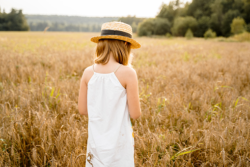 Happy girl walks in beautiful wheat field, embracing summer's yellow sun, nature freedom outdoors. White dress, straw hat, surrounded by rye, barley. Autumn harvest time rural scene.Own piece of land.