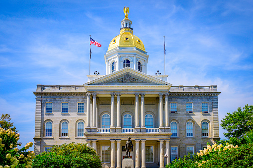 New Hampshire State House, Gold-domed State government office in Concord, New Hampshire, USA, American Flag and the state flag waving high in the wind over the green garden