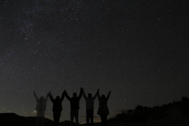 In this captivating image, silhouettes of people gather beneath an open gourd-shaped structure, set against a backdrop of a dark, starry night. The play of shadows against the night sky creates a striking visual contrast, offering a sense of wonder and mystery. The open gourd frames the scene, adding an intriguing element to this mesmerizing night setting.