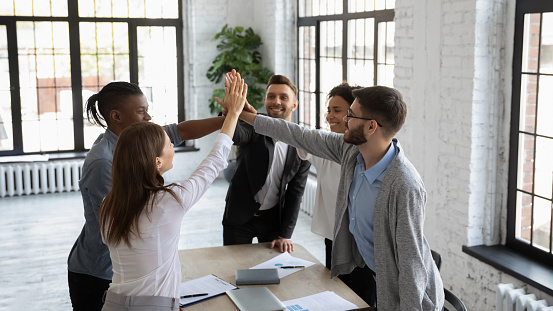 Excited multiracial colleagues give high five celebrate shared company group win or victory at office meeting, overjoyed diverse businesspeople join hands engaged in teambuilding activity at briefing