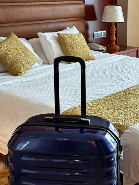istock Close-up image of blue wheelie suitcase besides double bed with leatherette headboard, stacked white pillows, gold cushions, folded duvet, electric lamp on nightstand, focus on foreground 1655532261