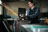 Young woman grinding a metal piece on vise with angle grinder in metal workshop