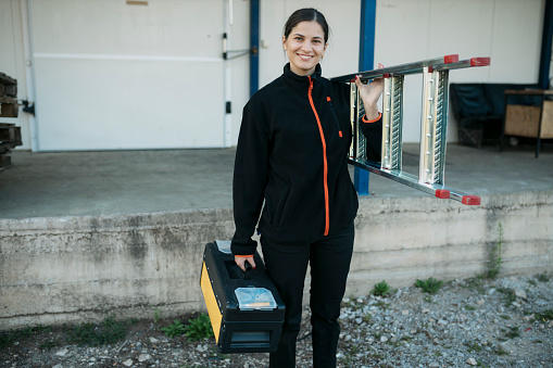 Portrait of a joyful professional builder holding a toolbox and ladder. Female worker standing outside with toolkit and ladder looking at camera and smiling.