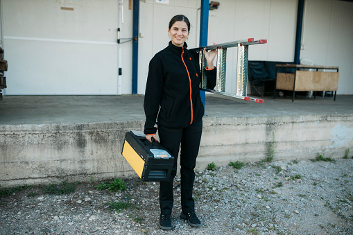 Full length portrait of a woman worker holding a tool box and ladder outside. Female construction contractor standing outside looking at camera and smiling.