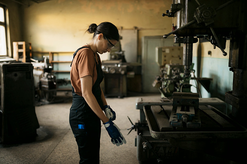 Side view of a female worker putting on safety gloves while standing at drill machine in metal workshop. Woman working in a small metal workshop.